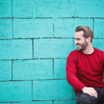 Surgery for puffy nipples in men: man wearing red sweatshirt and black pants leaning sitting in front of turquoise brick wall.