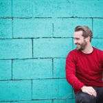 Surgery for puffy nipples in men: man wearing red sweatshirt and black pants leaning sitting in front of turquoise brick wall.