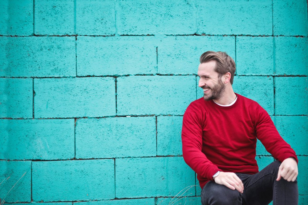 Surgery for puffy nipples in men: man wearing red sweatshirt and black pants leaning sitting in front of turquoise brick wall.